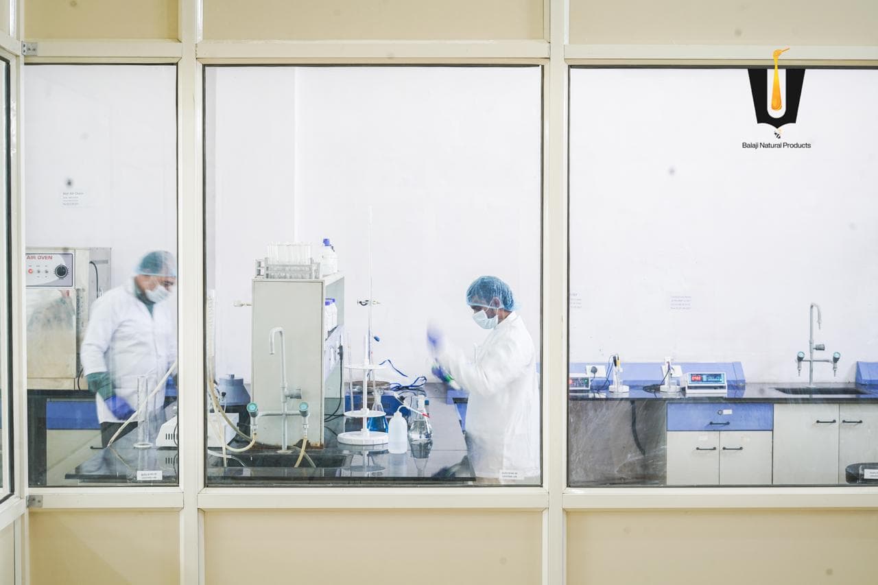 Two laboratory technicians in coats, hairnets, and masks testing honey samples with glassware in the Balaji quality lab