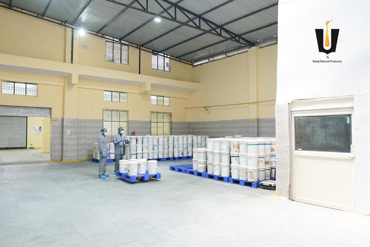 Workers in PPE moving white honey buckets on blue pallets with a pallet jack in the storage hall